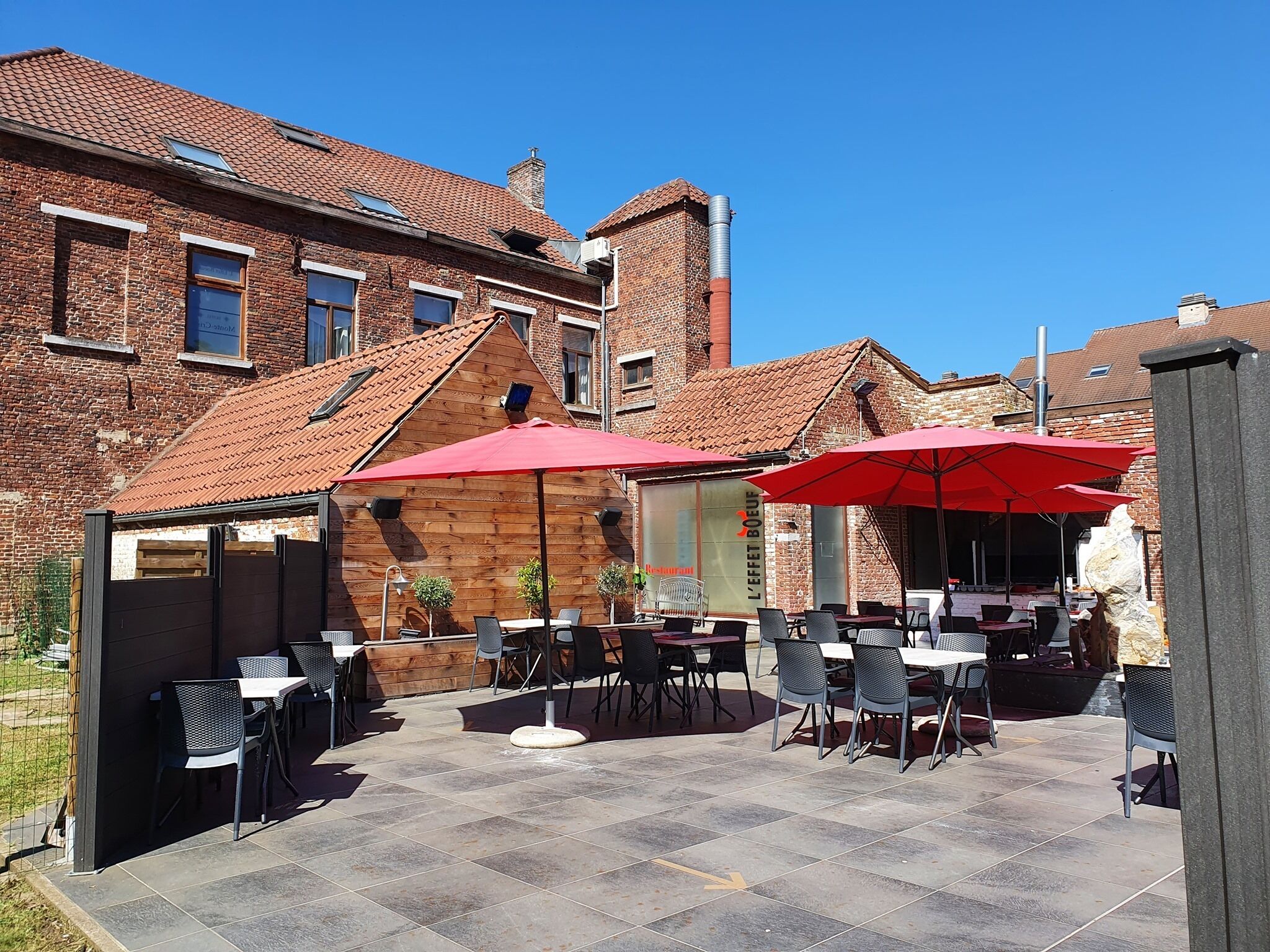 Una terrazza esterna con tavoli e sedie, ombrelloni rossi e un edificio in mattoni sullo sfondo.