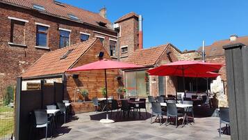 Una terrazza esterna con tavoli e sedie, ombrelloni rossi e un edificio in mattoni sullo sfondo.