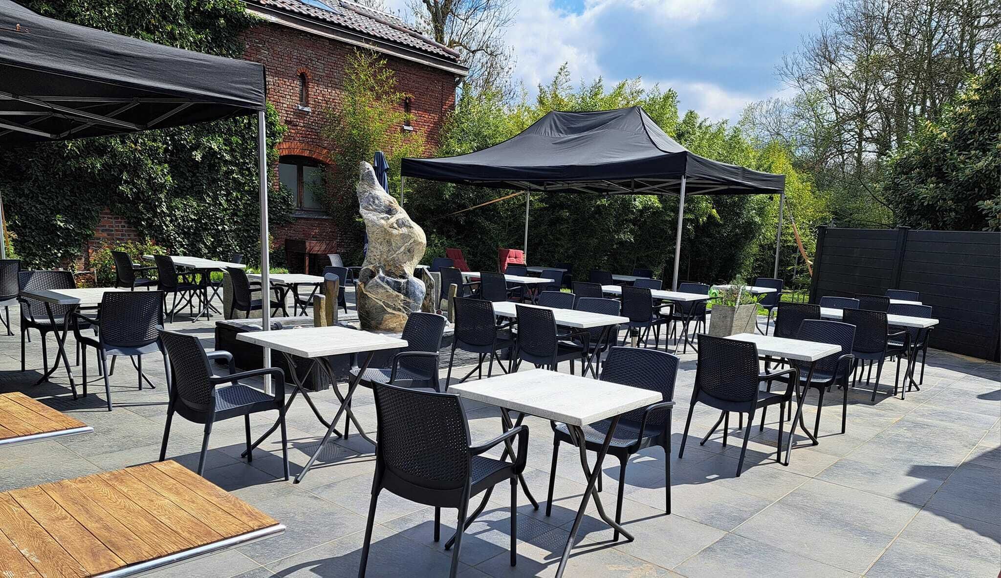 Eine Terrasse im Freien mit Tischen und Stühlen, eine große Steinskulptur und ein Backsteinhaus im Hintergrund.