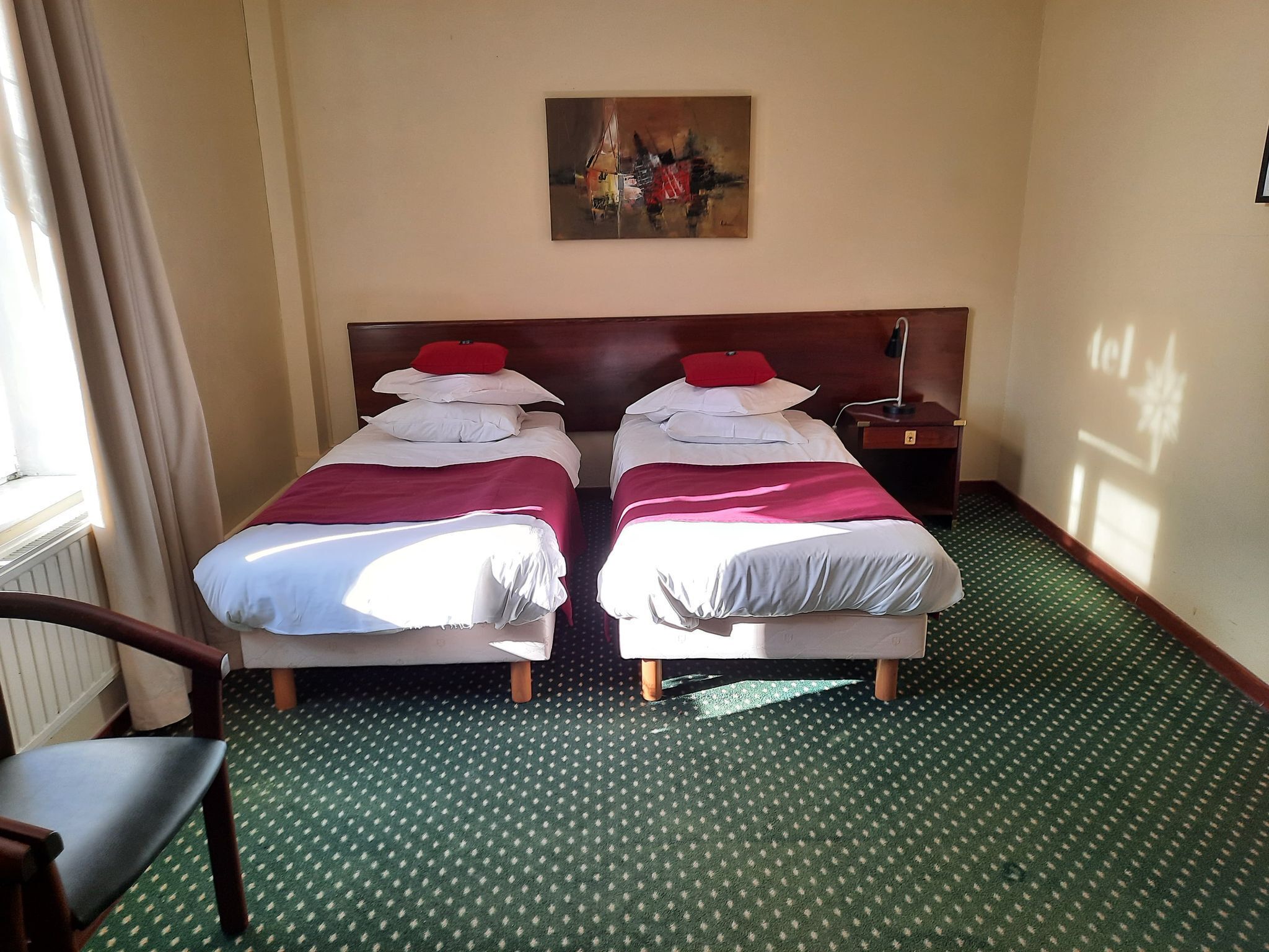 Habitación de hotel con dos camas, un cuadro en la pared, una silla y una mesita de noche con lámpara.