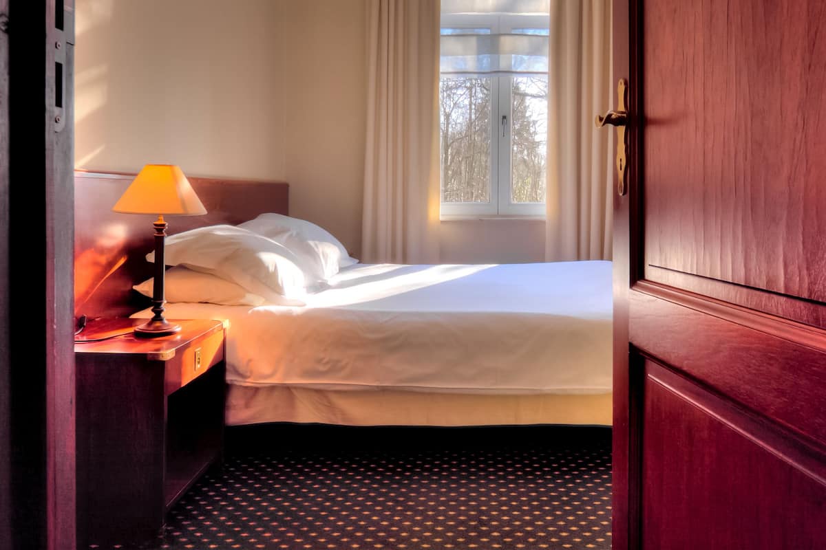A hotel room with a bed, a nightstand with a lamp, a window with curtains, and a wooden door.
