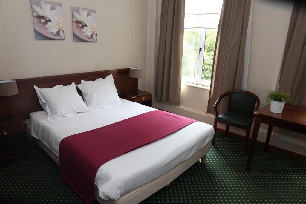 A hotel room with a large bed, two pillows, a desk with a chair, a small table with a plant, and a window with curtains.