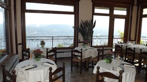 A dining area with tables set for two, large windows offering a view of a body of water and mountains, and a balcony with a metal railing. - Hotel Panoramico lago d'Orta adults only (Madonna del Sasso)