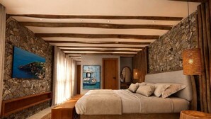 A bedroom with a stone wall, wooden beams, a bed, a nightstand, and a woven rug. - Margaridas Boutique Hotel - antiga Pousada Jardim das Margaridas (Porto Seguro)