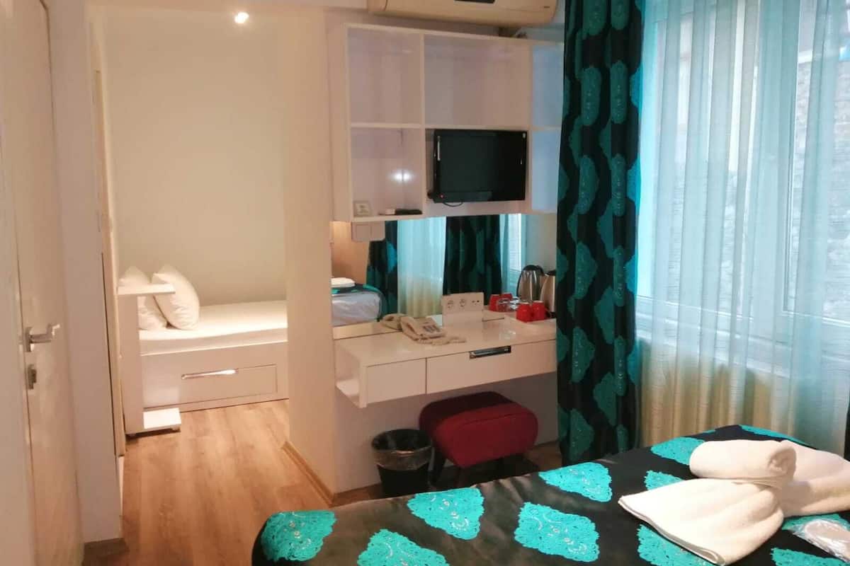 A compact hotel room with a bed, a desk with a computer, a TV, and a window with curtains.