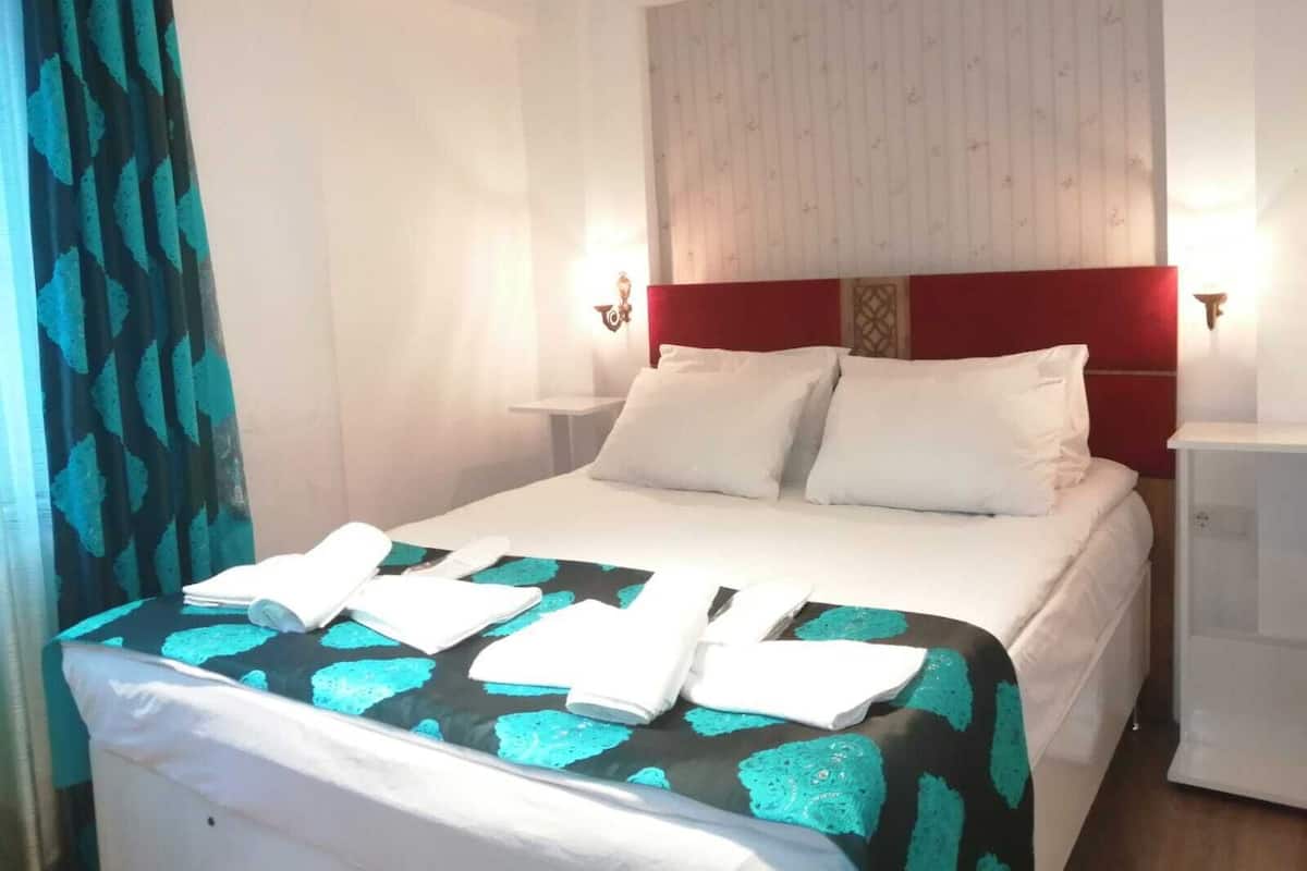 A hotel room with a bed, white pillows, a red headboard, and a nightstand with a lamp.