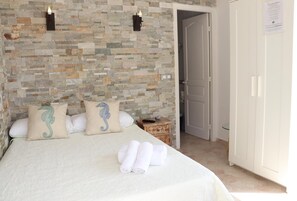 A neatly made bed with a white bedspread and two decorative pillows, a nightstand with a remote control, and a stone-patterned wall. - Hostal La Paloma I (Calpe)