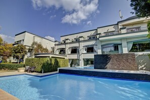Un espace piscine moderne avec une piscine à l’eau d’un bleu limpide, un coin salon avec des chaises, et un bâtiment doté de nombreux balcons et de grandes fenêtres.