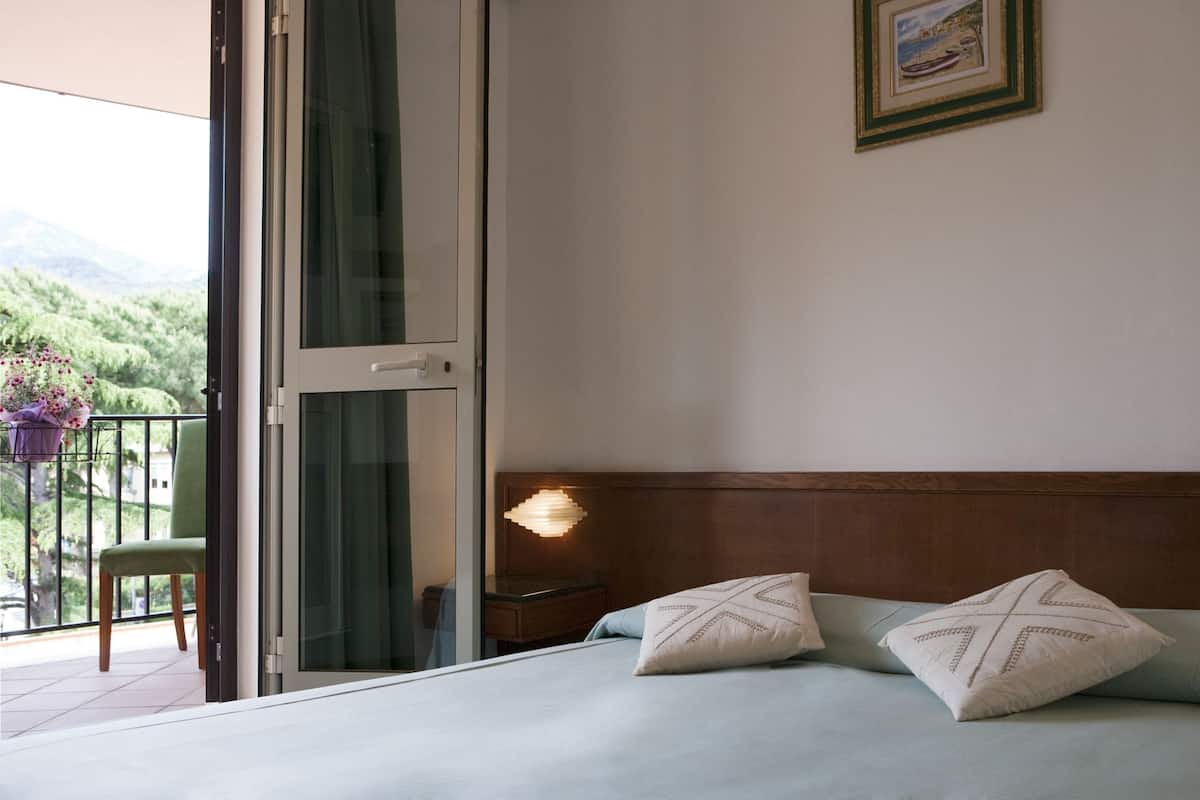 A bedroom with a bed, pillows, a nightstand, a lamp, a chair, and a balcony with a view of greenery.