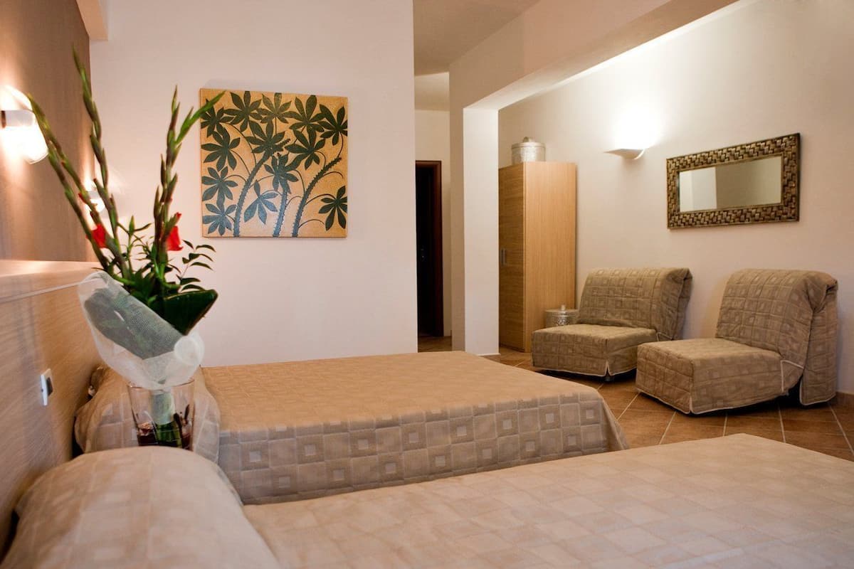 A hotel room with two beds, a sitting area with two chairs, a wall-mounted mirror, and a painting of tropical foliage.