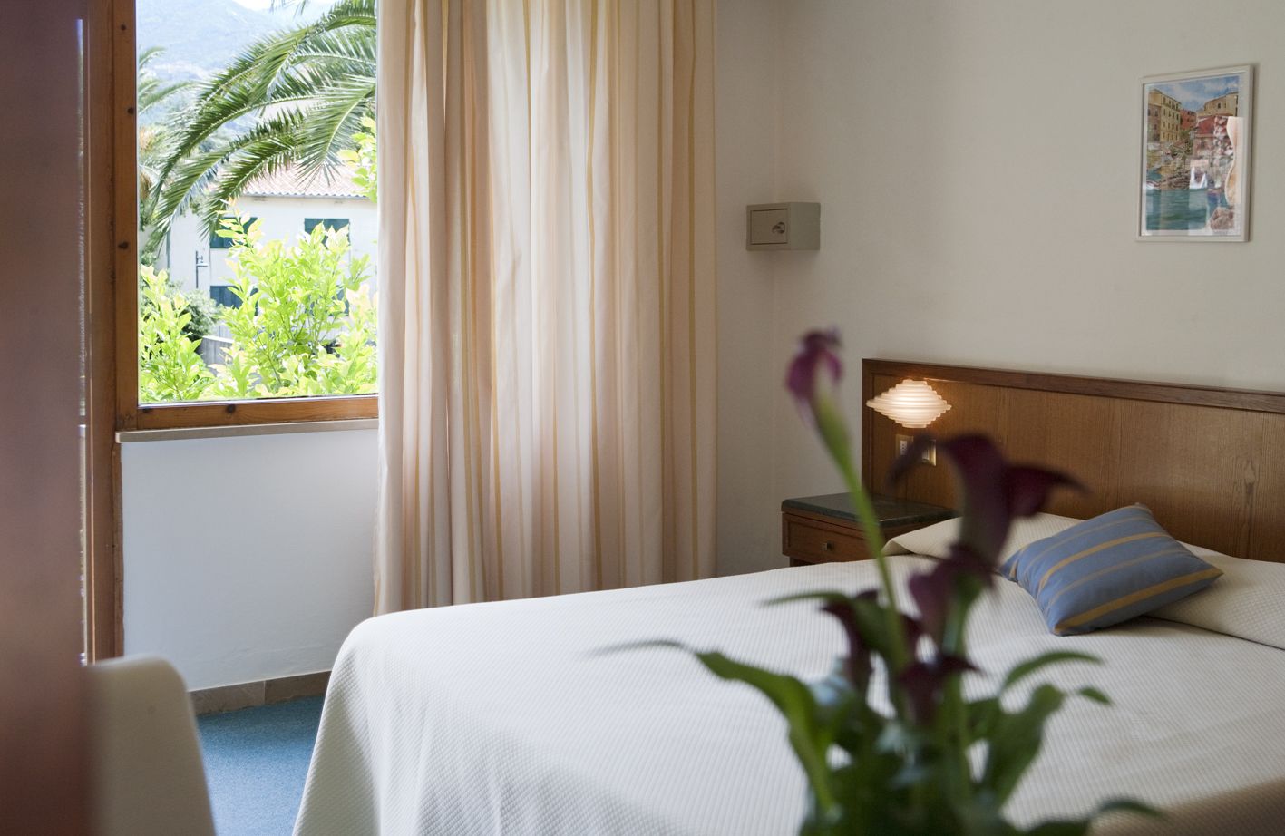 Una camera d'albergo con un letto, un comodino, una lampada, una finestra con tende e vista su del verde.