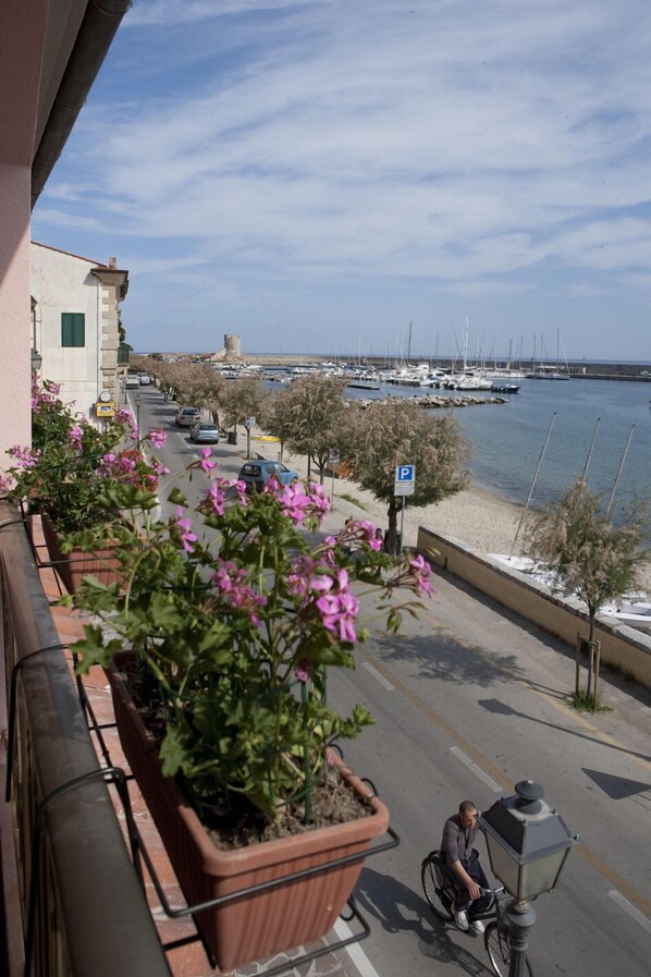 A balcony with potted plants, a street with parked cars, and a view of boats on the water.