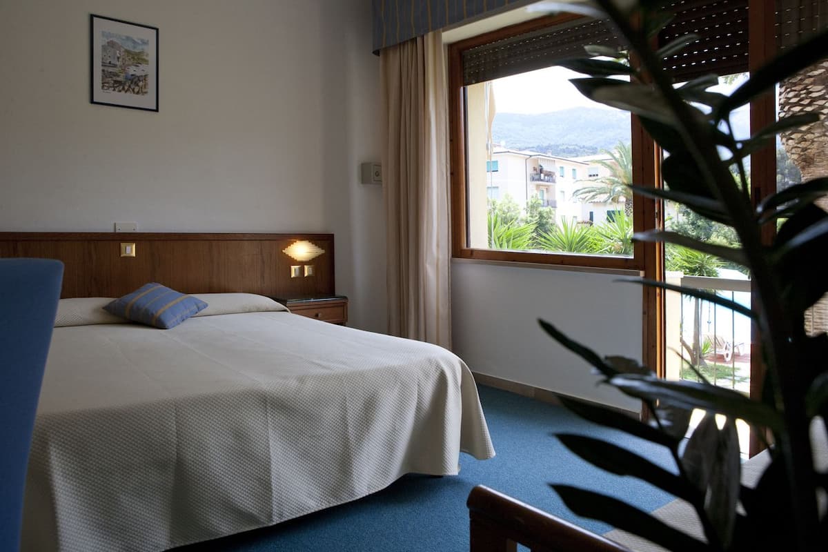 A hotel room with a bed, a nightstand, a lamp, a window with a view of buildings and greenery, and a plant in the foreground.