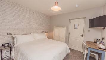 Een slaapkamer met een wand in baksteenmotief, een bed met wit beddengoed, een bureau met stoel en een aan de muur bevestigde televisie.