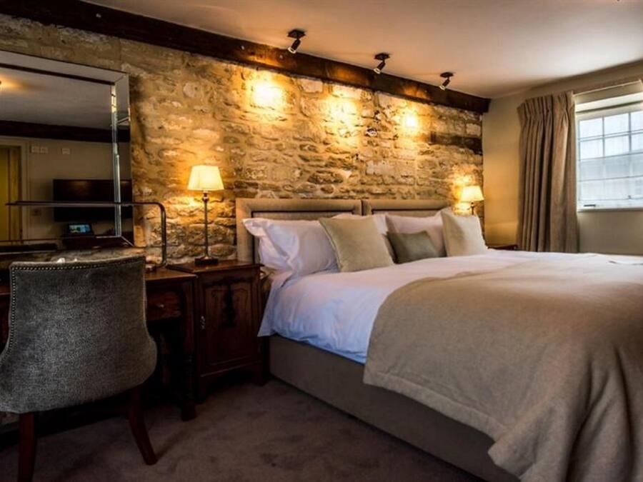 A bedroom with a stone wall, a bed with white linens, a desk with a chair, and two bedside lamps.