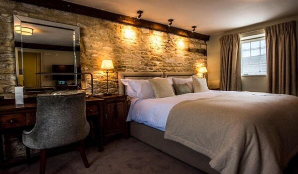 A bedroom with a stone wall, a bed with white linens, a desk with a chair, and two bedside lamps.