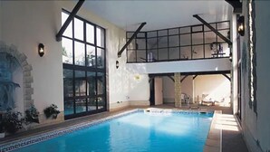 Piscina interior con vistas al cielo a través de amplios ventanales, terraza superior cubierta y pared de piedra con plantas.