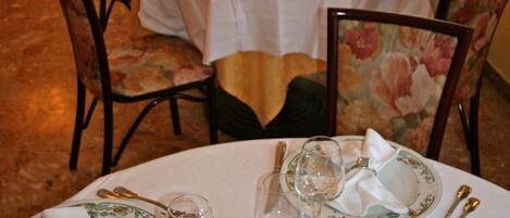 A dining area with round tables set for a meal, each with a white tablecloth, floral chairs, and empty wine glasses.