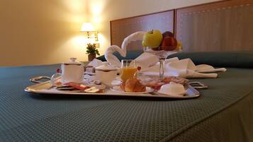 Ein Frühstücksbrett mit Croissants, Orangensaft in einem Glas, einem Teekännchen und einer Obstschale auf einem Bett.