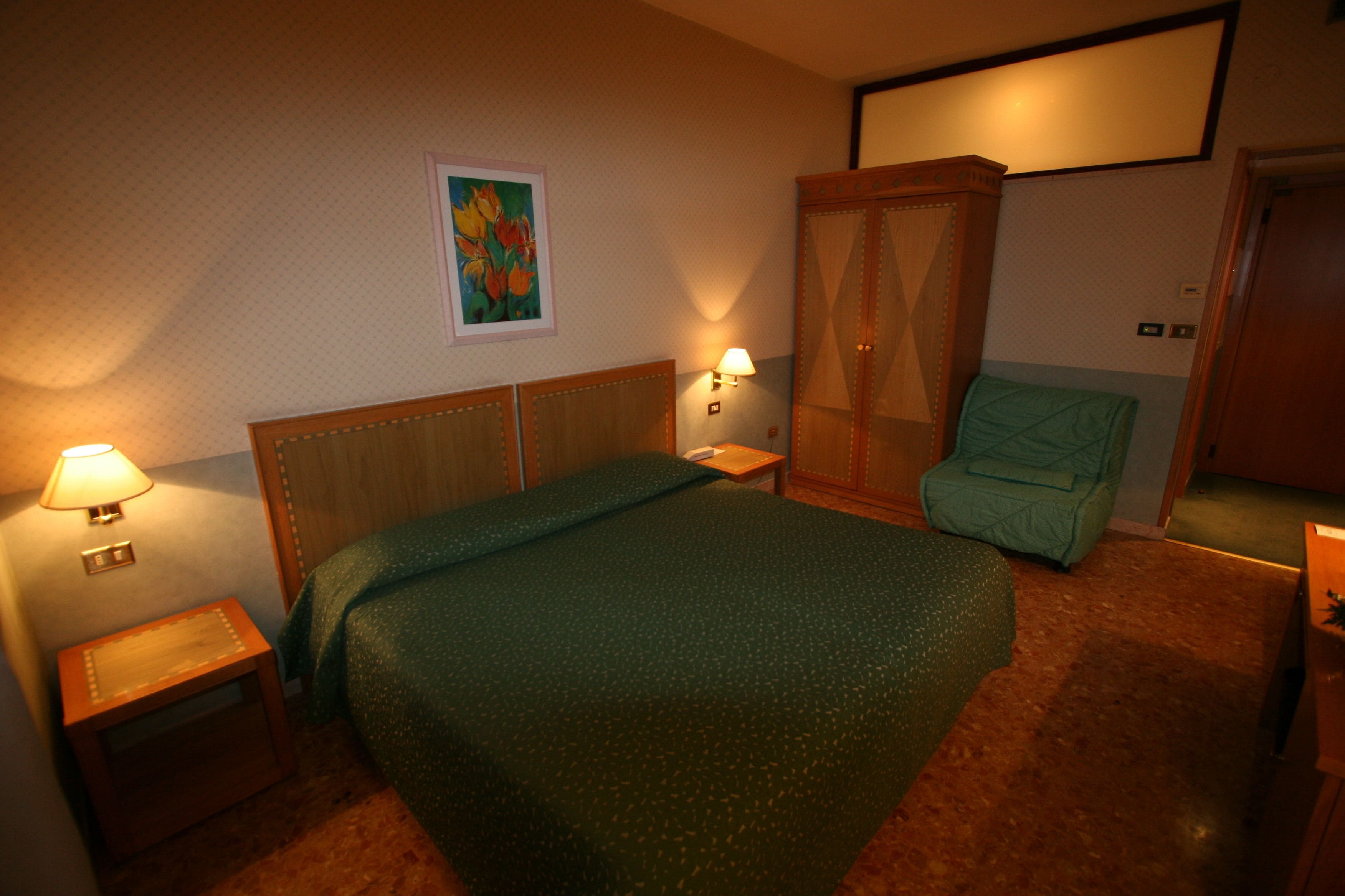 Una camera d'albergo con un letto, una poltrona verde, un armadio e due lampade ai comodini.