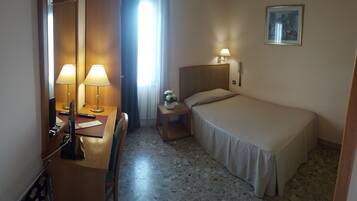 Una camera d'albergo con un letto, un comodino con una lampada, una scrivania con lampada, una sedia, un telefono e un quadro appeso al muro.