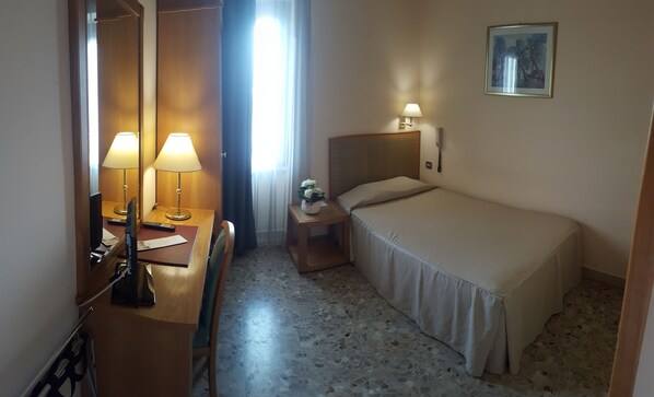 Habitación de hotel con cama, mesita de noche con lámpara, escritorio con lámpara, una silla, teléfono y un cuadro en la pared.