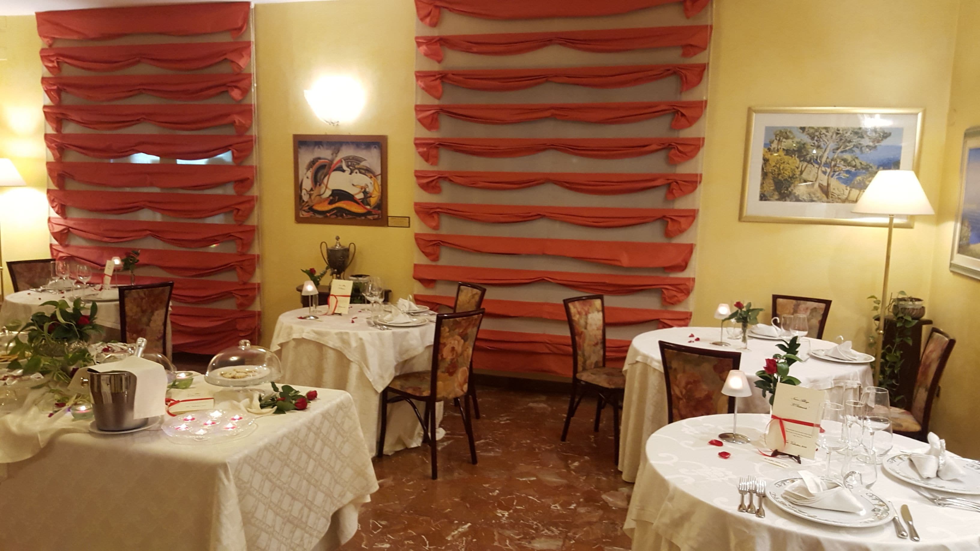 Un espace repas avec des tables dressées pour le service, des rideaux rouges aux murs et un tableau accroché au-dessus des tables.