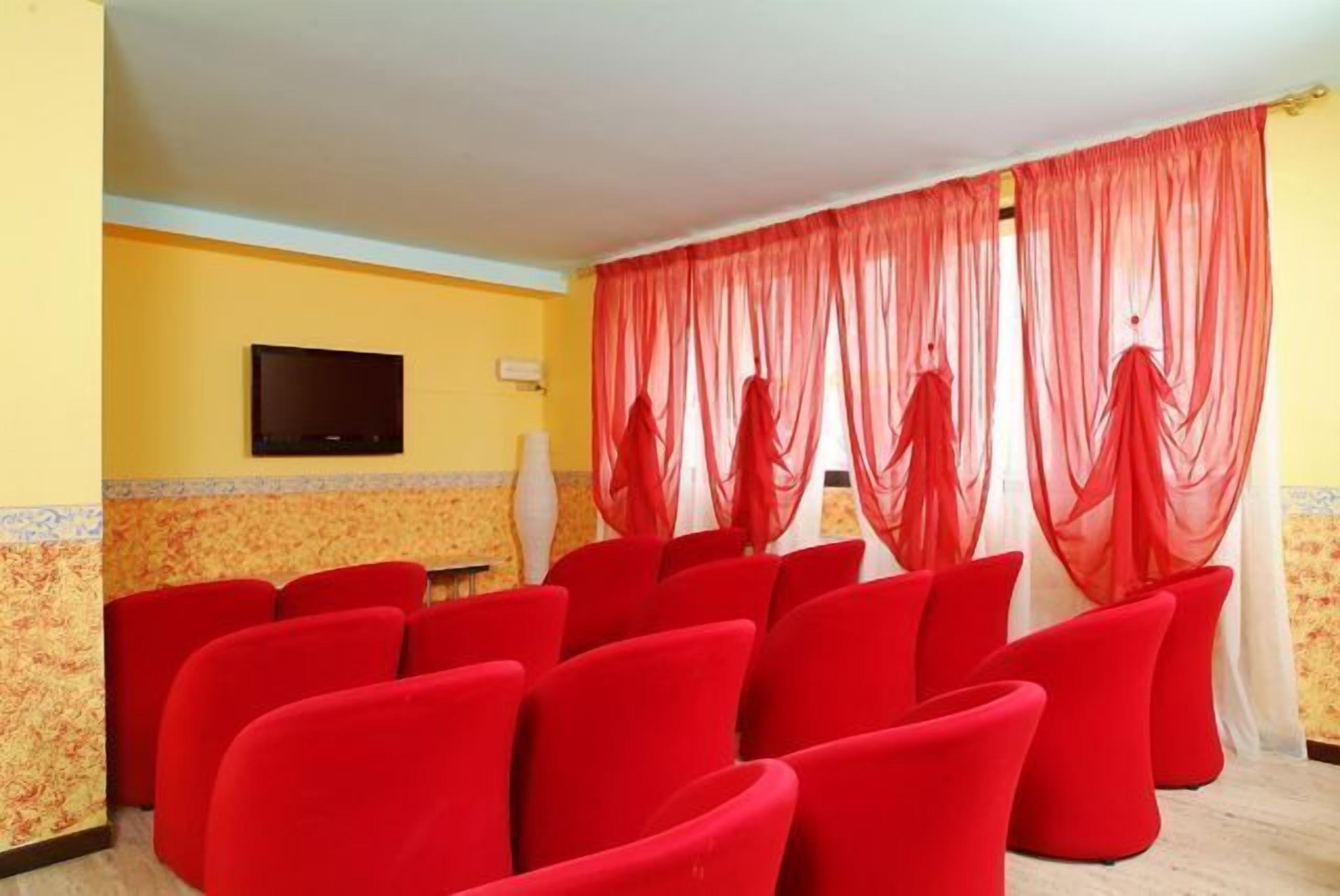 A room with red chairs arranged in rows, a flat-screen TV mounted on the wall, and sheer red curtains.