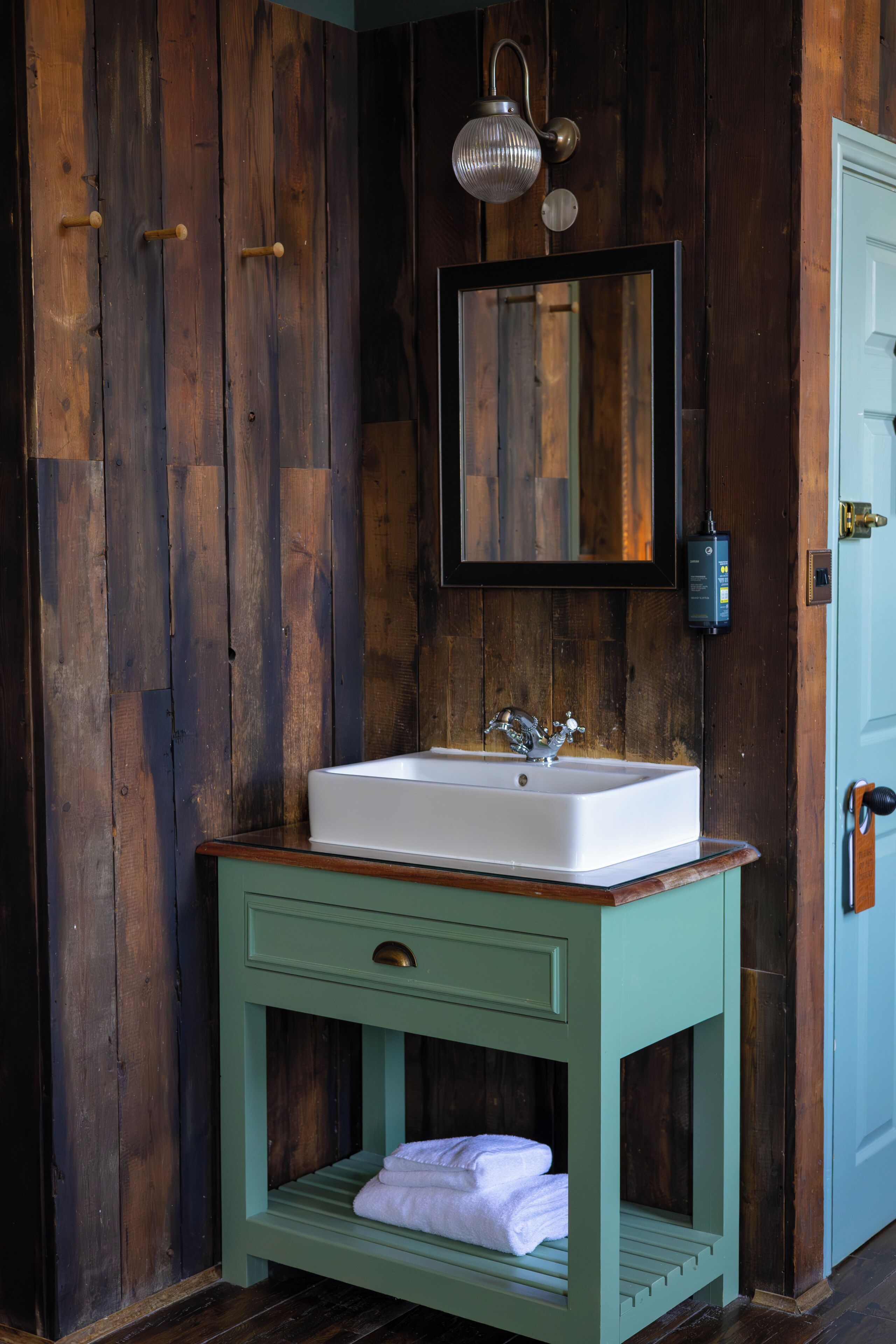 A bathroom with a wooden wall, a white sink, a green vanity, and a hanging light fixture.