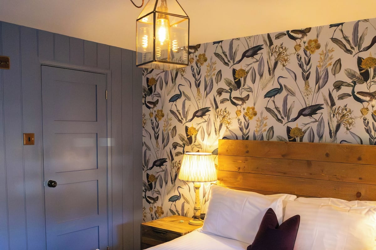 A hotel room with a bed, a wooden headboard, a wall with floral wallpaper, a door, and a nightstand with a lamp.