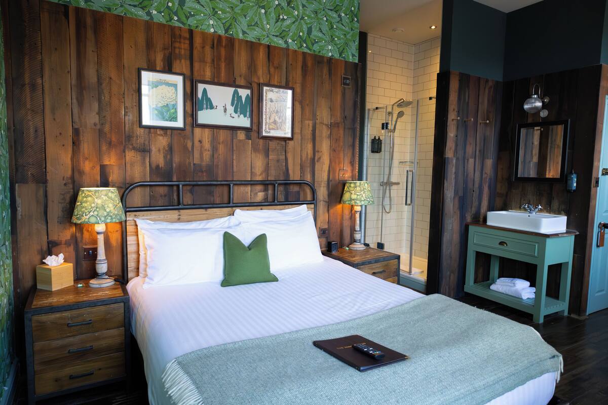 A hotel room with a wooden headboard, a bed with white linens, a green pillow, a bedside table with a lamp, a nightstand, a bathroom with a shower, and a sink.