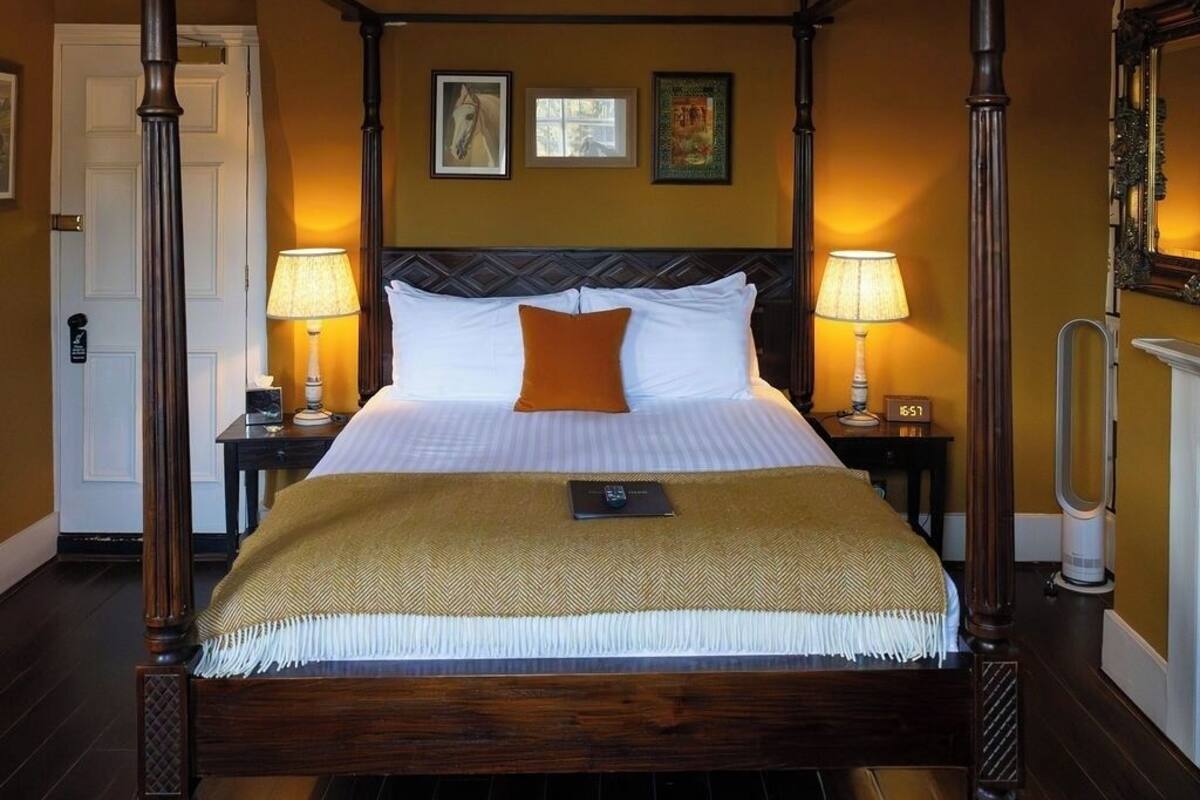 A four-poster bed with a headboard, two bedside tables with lamps, a nightstand, a mirror, and a door.