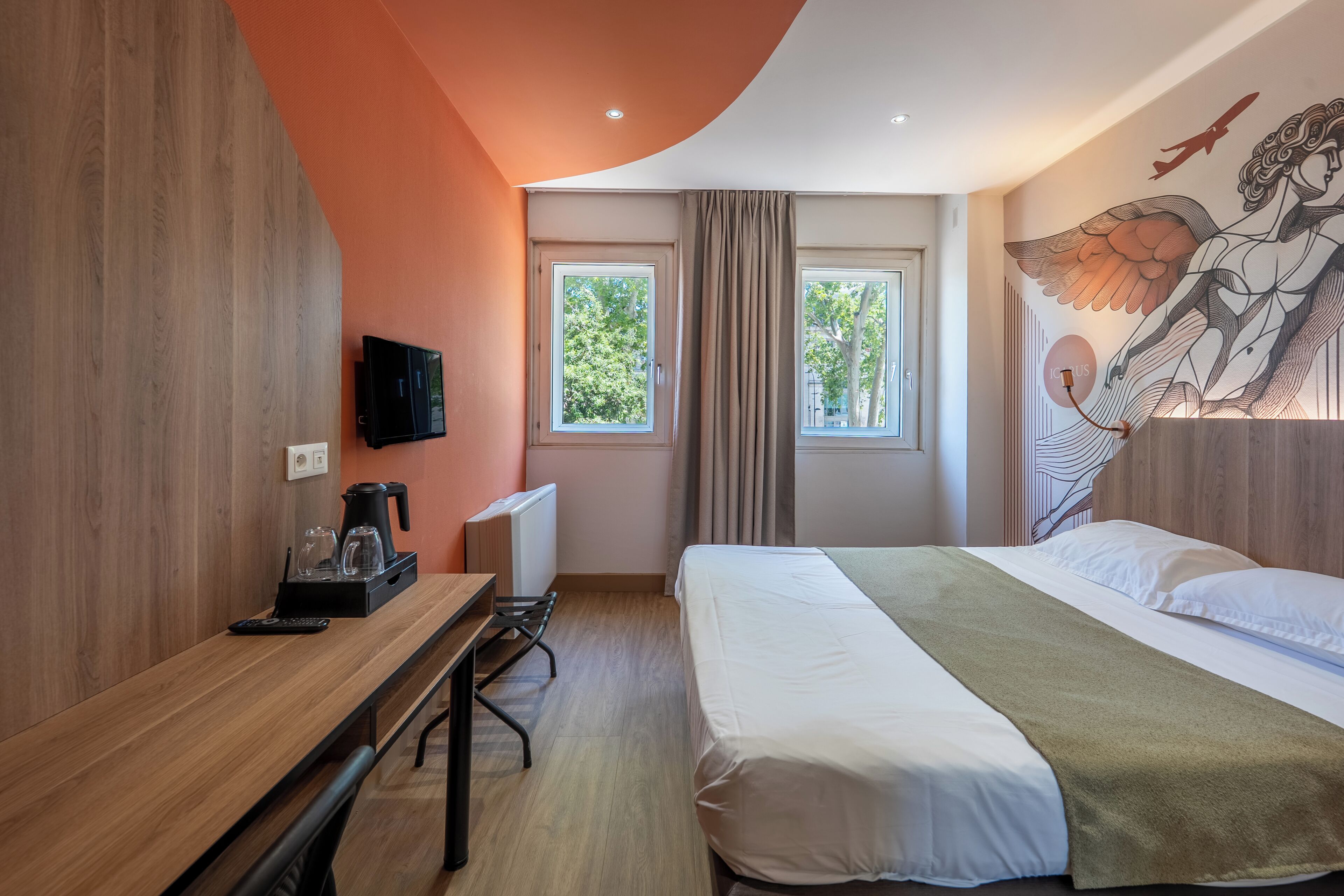 Ett modernt hotellrum med ett skrivbord i trä, en säng med ett grönt överkast, en tavla på väggen och ett fönster med gardiner.