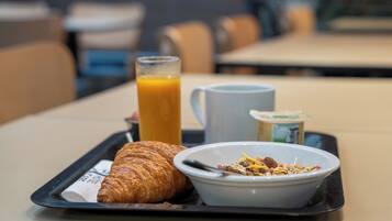 Ein Frühstücksbrett mit einem Croissant, einem Glas Orangensaft, einer Tasse und einer Schüssel Müsli mit Früchten und Nüssen.