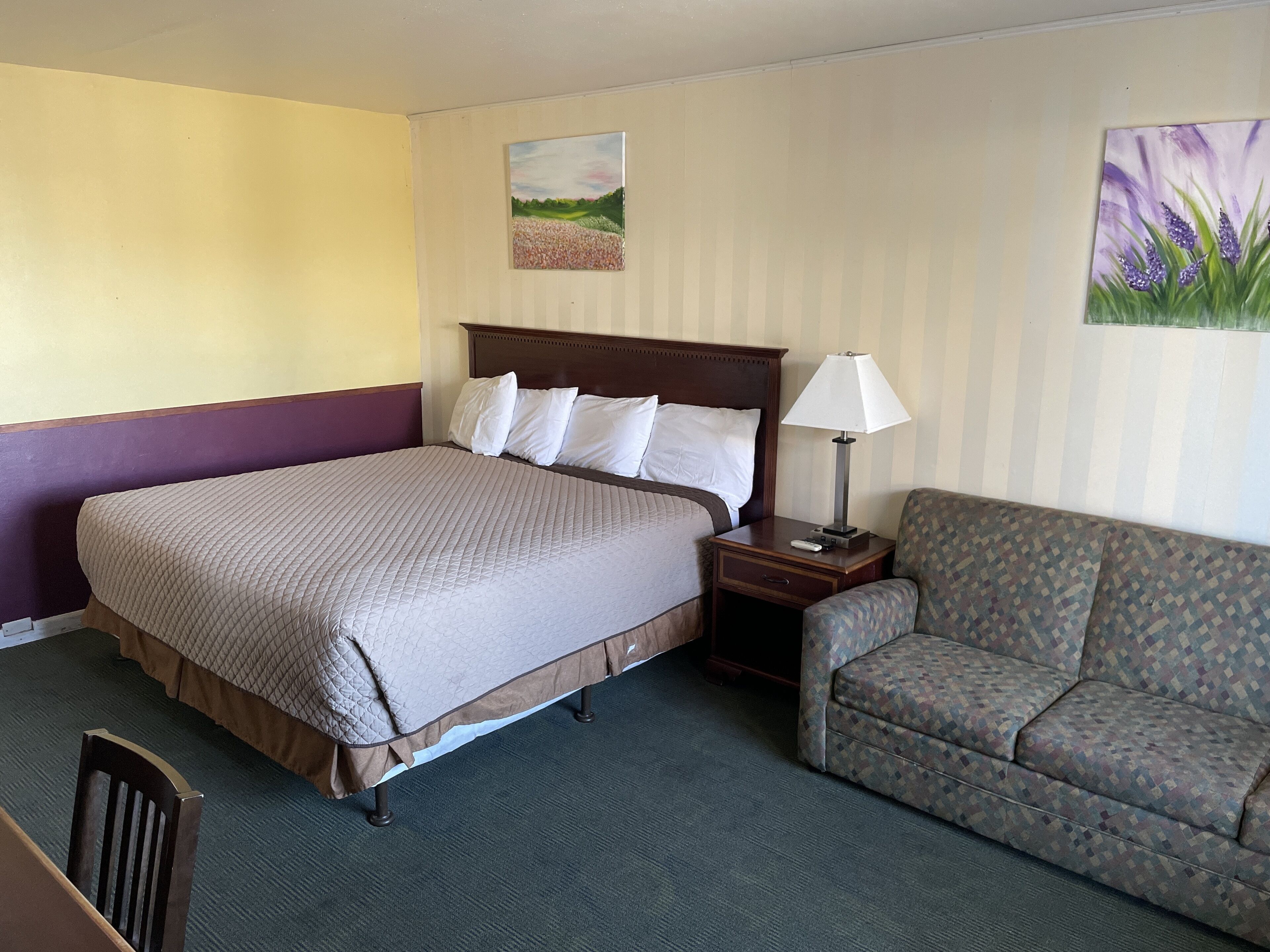 A hotel room with a bed, a nightstand, a lamp, a sofa, and two paintings on the walls.