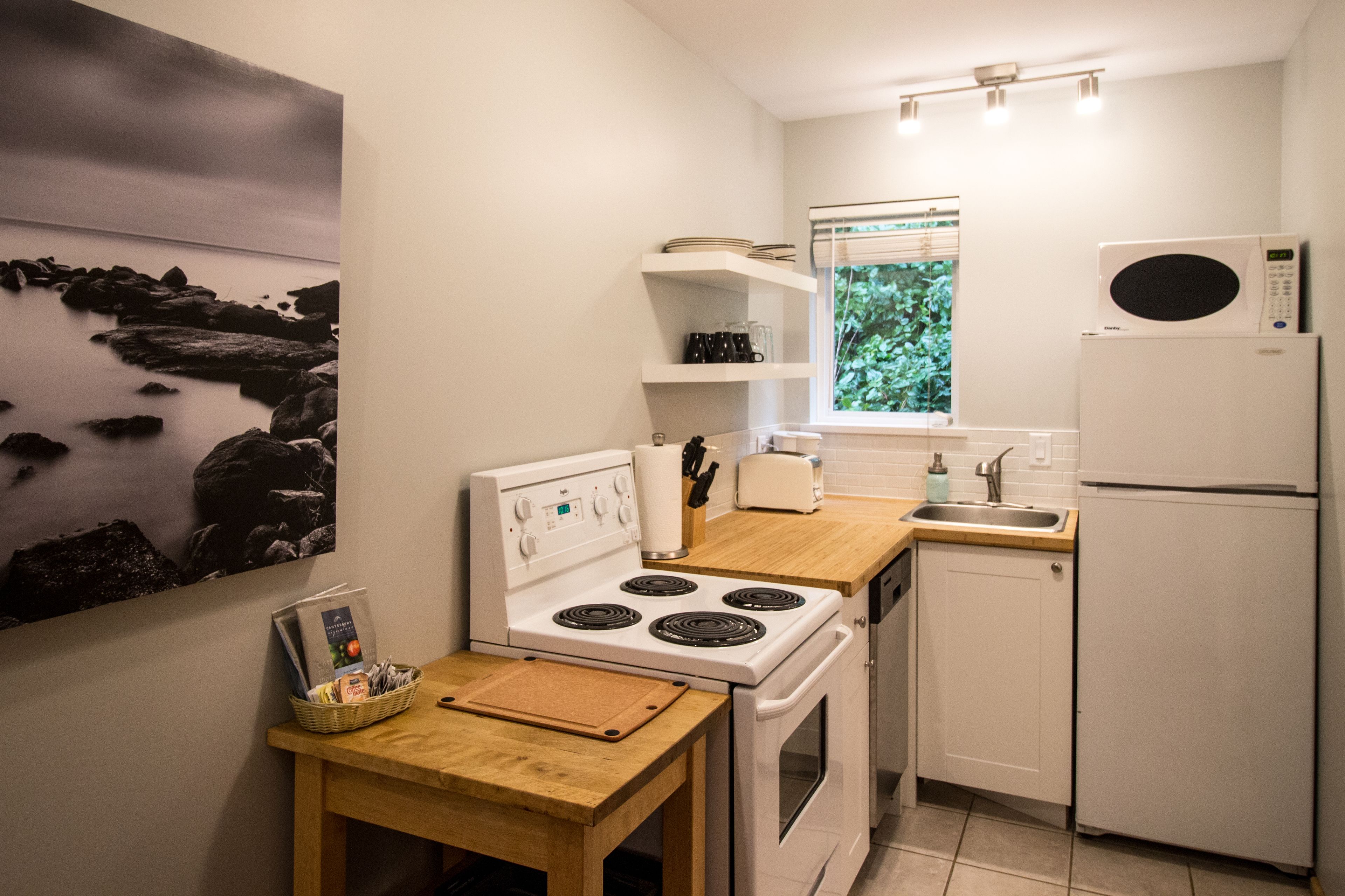 A compact kitchen with white appliances, a wooden table, and a window with a view of greenery.