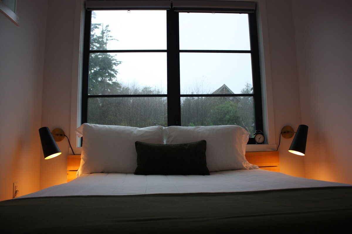 Quarto com uma janela grande, cama com roupa de cama branca, duas luminárias de cabeceira e um relógio na parede.