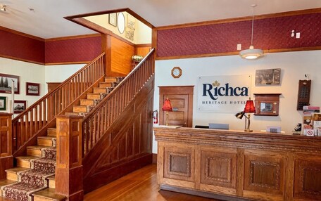 A hotel lobby with a wooden staircase, a reception desk, and a sign reading 'Ricchea Heritage Hotel'.