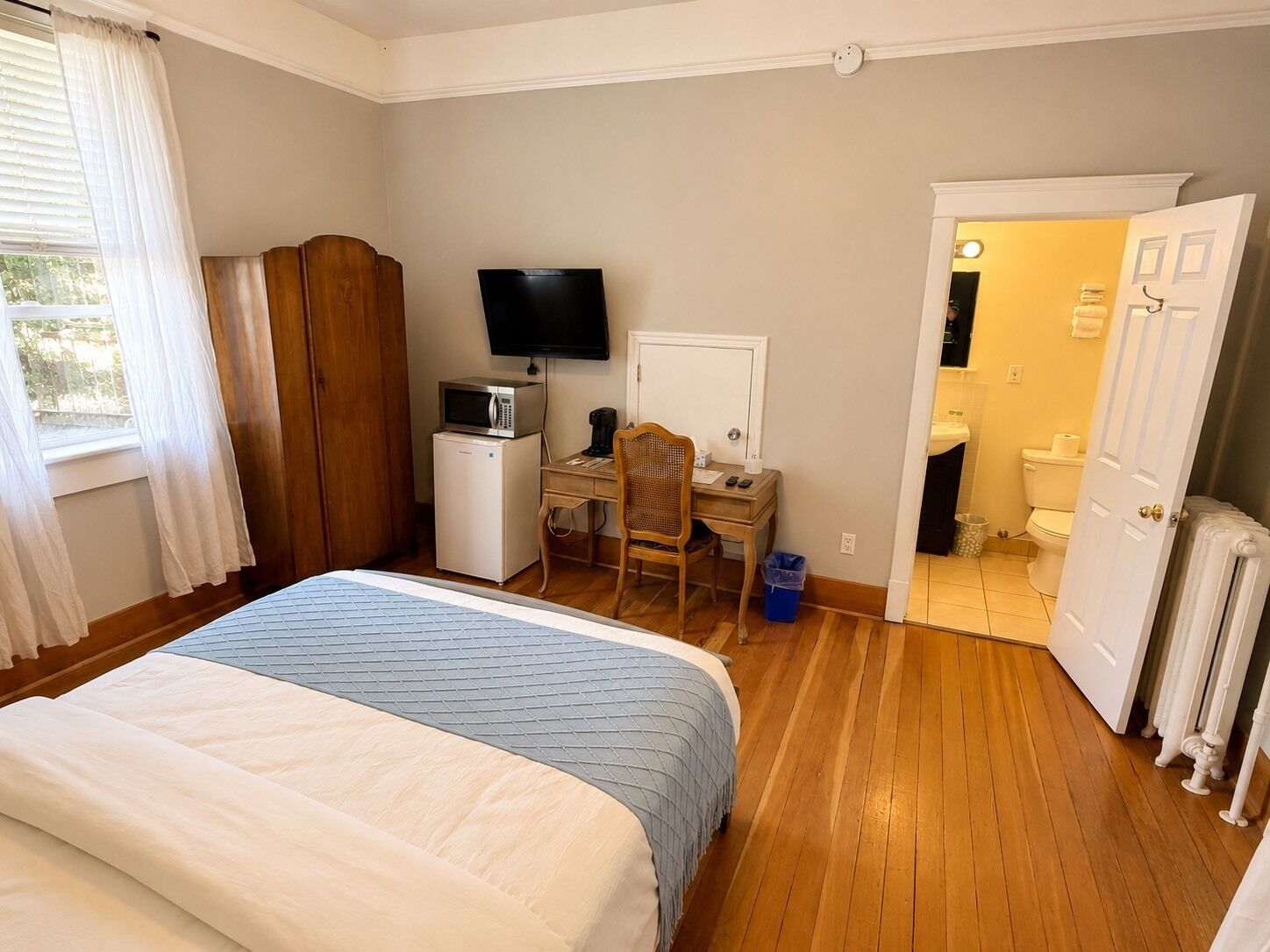 Un dormitorio con cama, escritorio con computadora, televisor y baño visible a través de una puerta abierta.