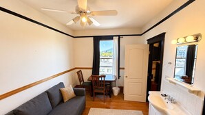 A compact room with a sofa, a dining table with chairs, a ceiling fan, a bathroom with a sink and mirror, and a view through a window. - Richca Heritage Hotel (Chilliwack)