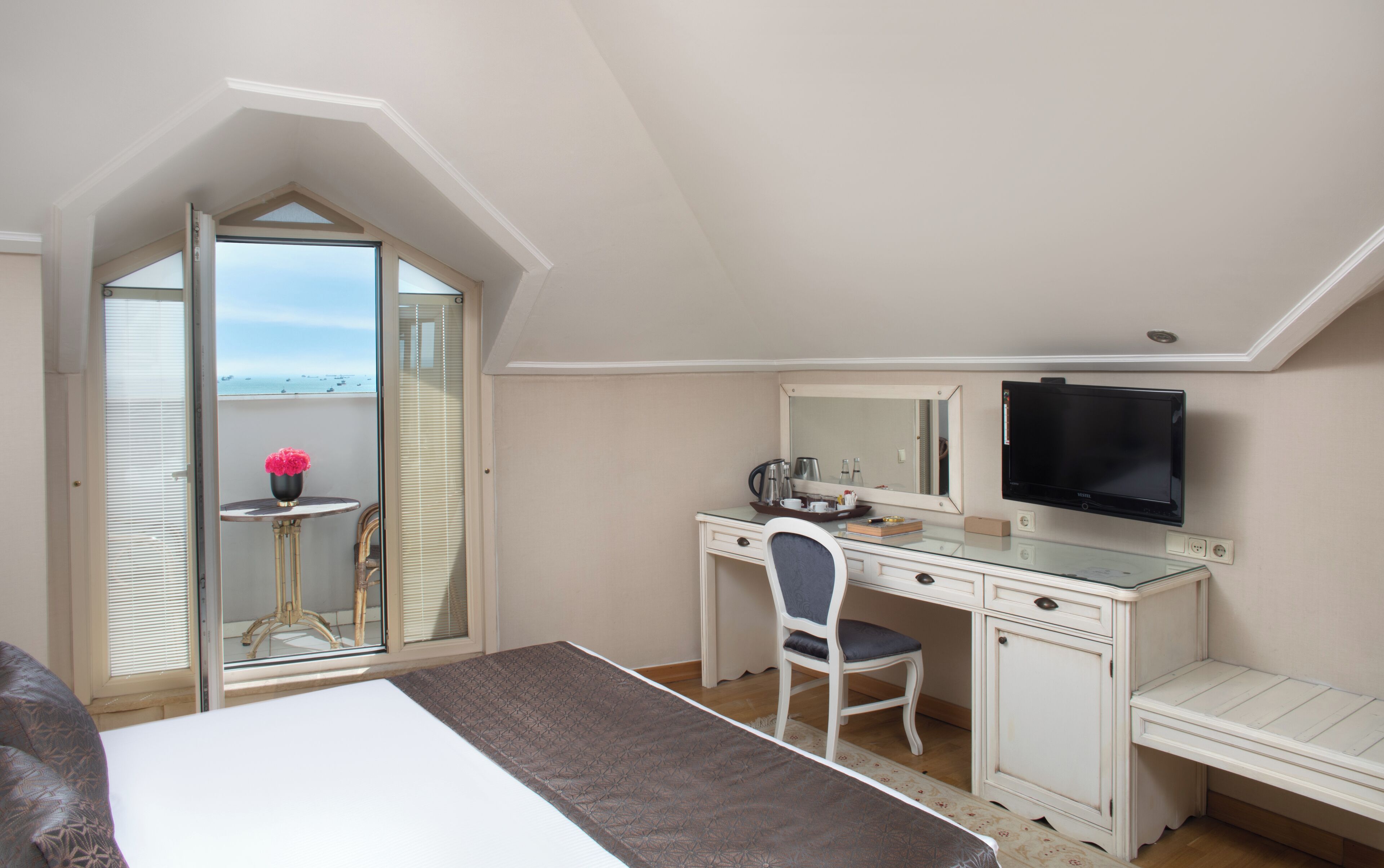 A bedroom with a bed, a desk, a chair, a television, and a balcony with a view of the sea.