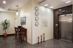 Un vestíbulo con mostrador de recepción, relojes, una planta en maceta y un ascensor.
