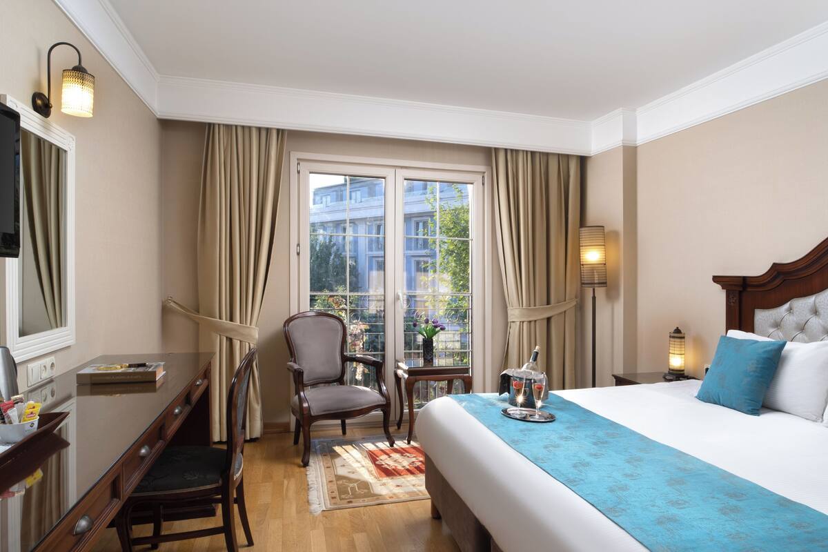 Une chambre d’hôtel comprenant un lit, un bureau, une chaise, une fenêtre avec des rideaux et une table de chevet avec une lampe.