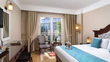 A hotel room with a bed, a desk, a chair, a window with curtains, and a nightstand with a lamp.