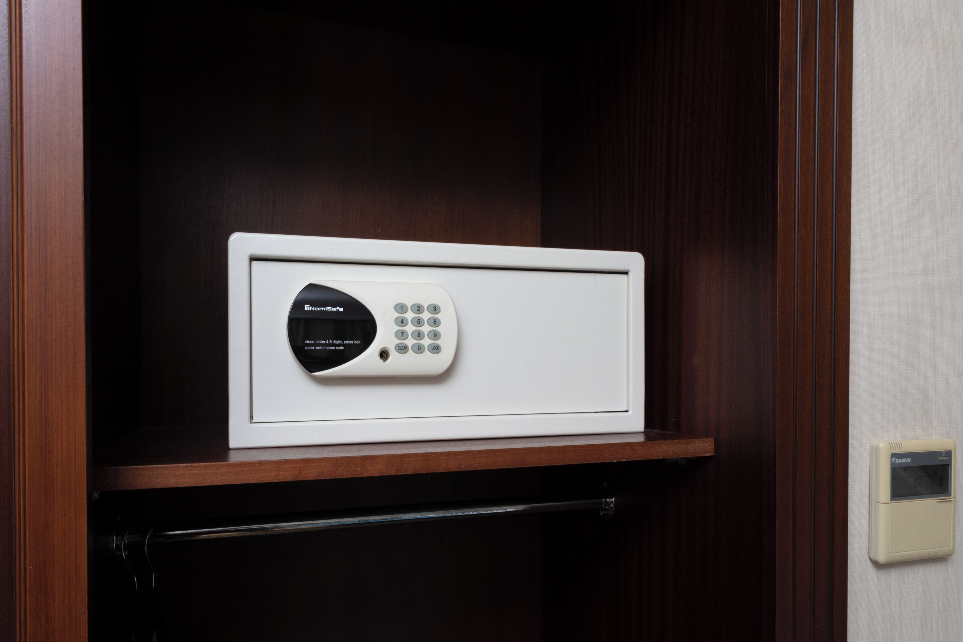 Un dispositivo electrónico blanco con teclado y pantalla, colocado sobre un estante de madera en una habitación con una puerta de madera oscura y un interruptor de luz en la pared.