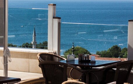 A seaside terrace with wicker chairs and a table, overlooking the ocean with a ship in the distance.