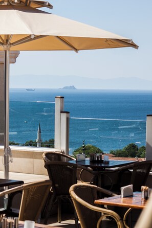 Una terraza frente al mar con sillones de mimbre y una mesa, con vista al océano y un barco a lo lejos.