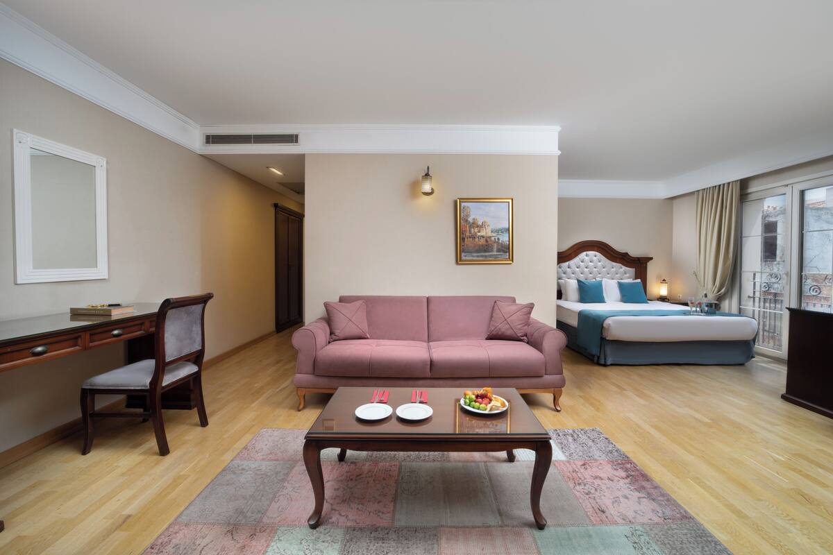 Une pièce avec un canapé rose, un bureau en bois, une table à manger avec des assiettes et un panier de fruits, un lit avec une literie bleue, et un tableau accroché au mur.