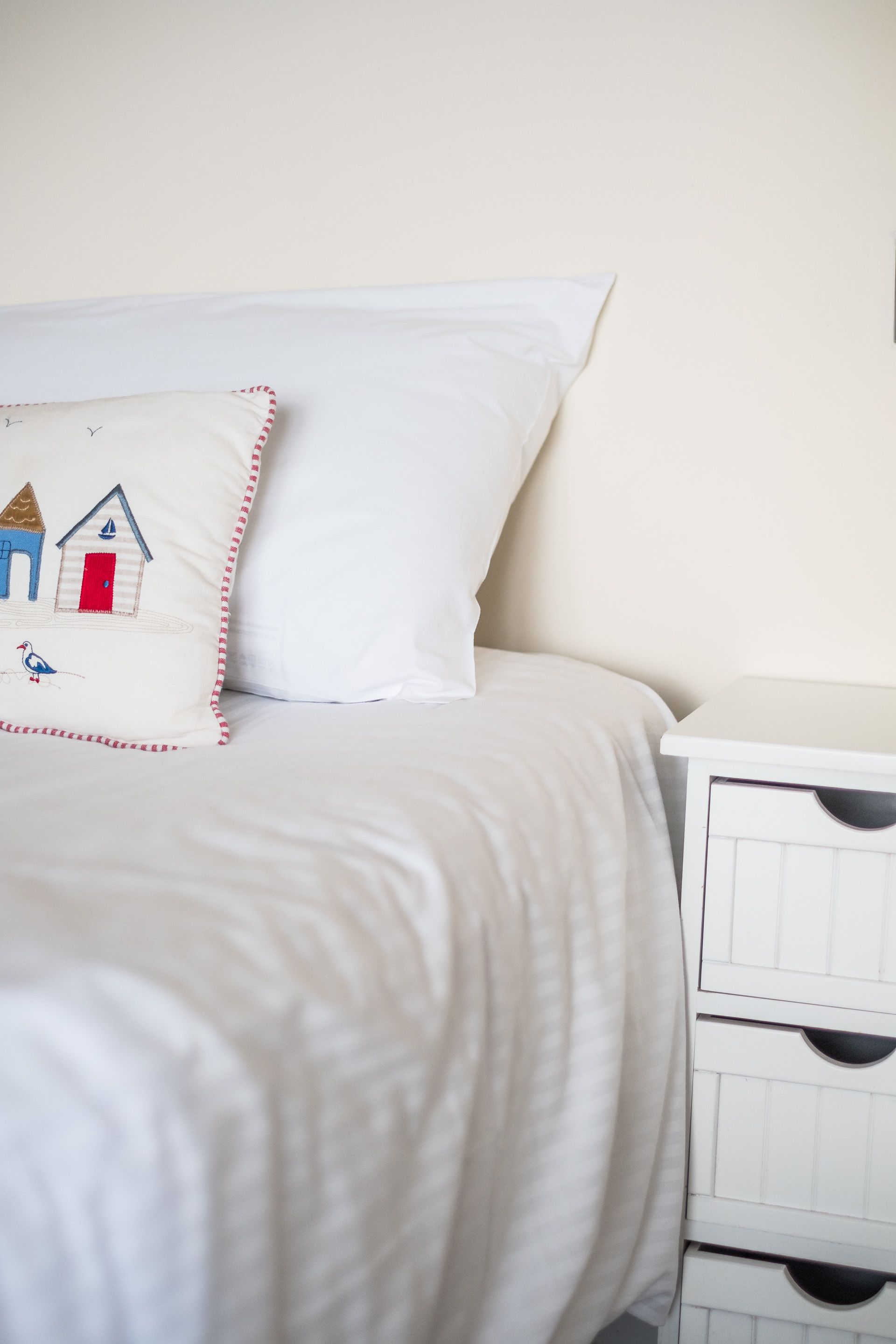 A neatly made bed with white bedding and a pillow featuring a seaside scene.