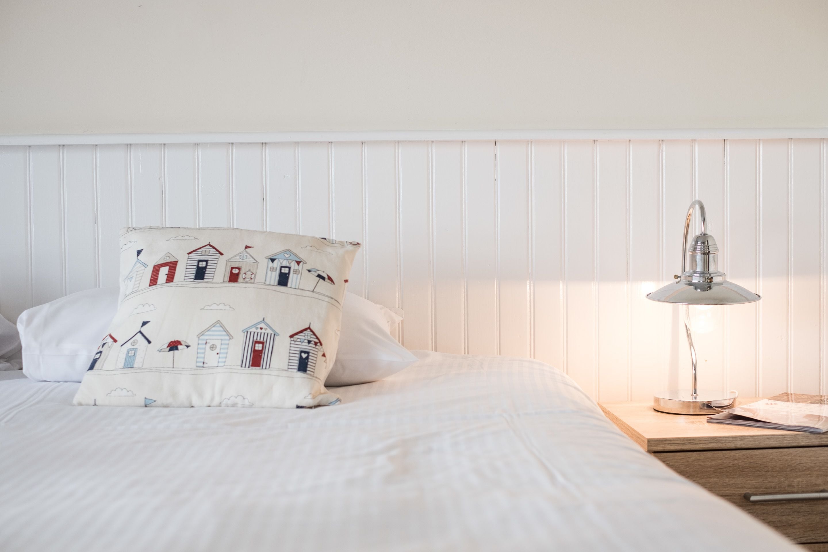 A neatly made bed with a white pillow featuring a seaside design, a bedside lamp, and a wooden nightstand.